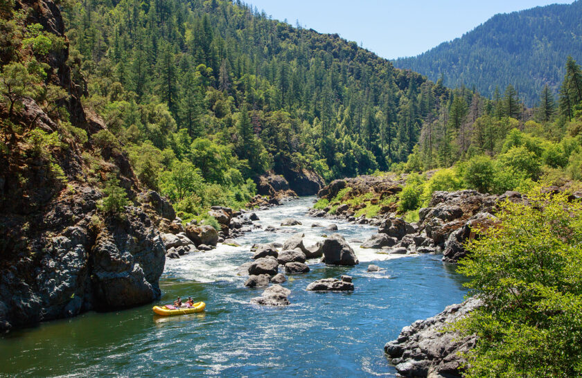 Scenic photo of a yellow raft entering Blossom Bar Rapid in a lush, forested valley on the Rogue River