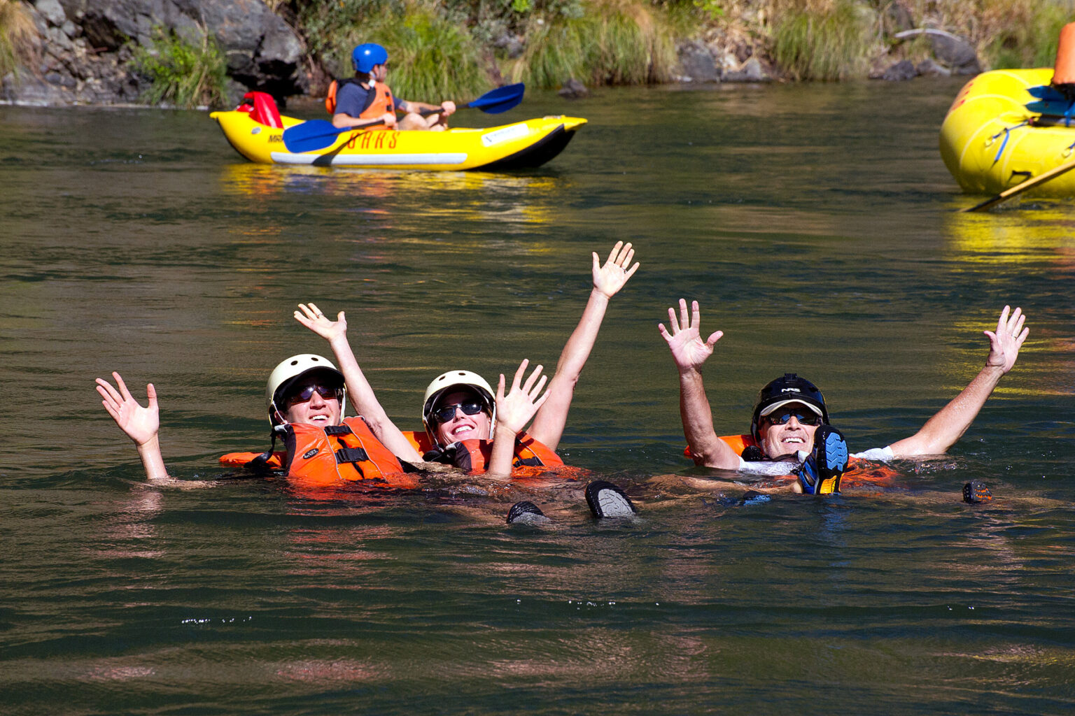 Three swimmers wave to the camera in calm waters of the Rogue River