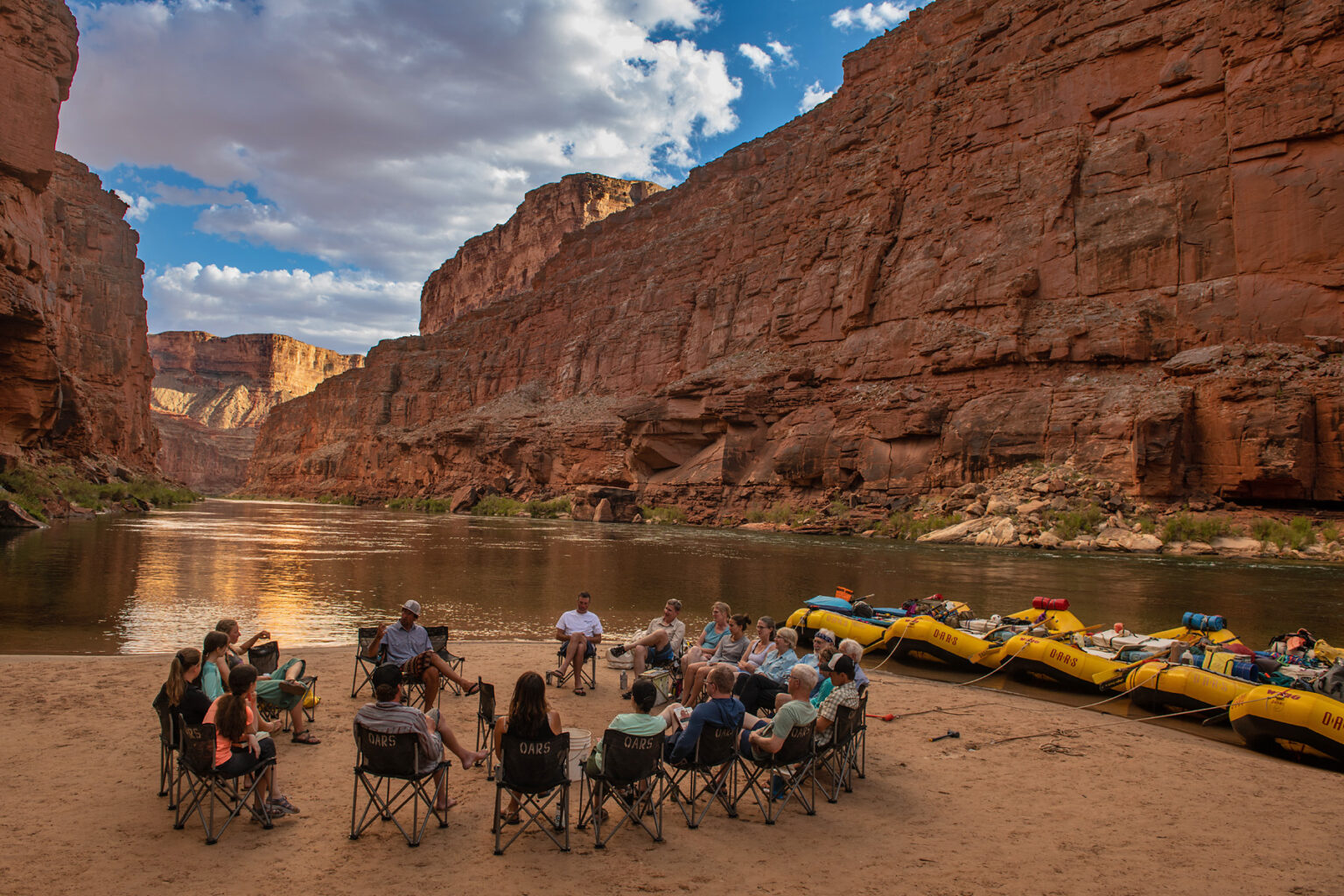 guides and guests in a chair circle on a sandy beach in Grand Canyon with five OARS rafts docked and staked for the night