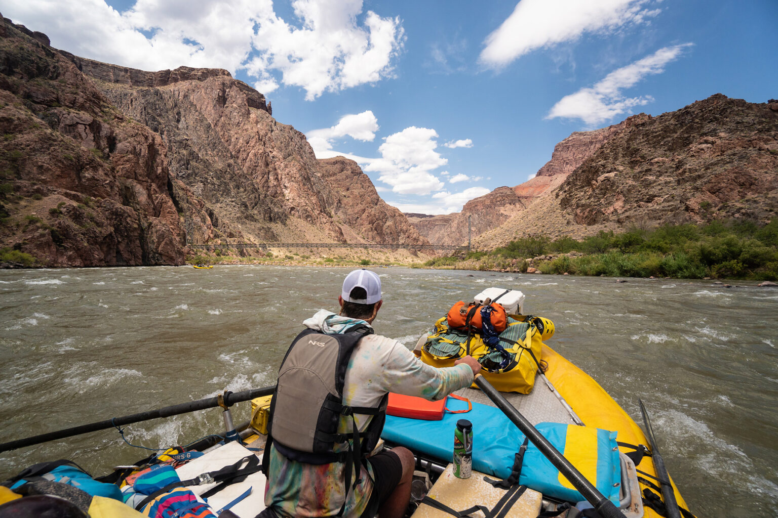 OARS rafting guide steers his raft down the Colorado River with a foot bridge to Phantom Ranch in the distance