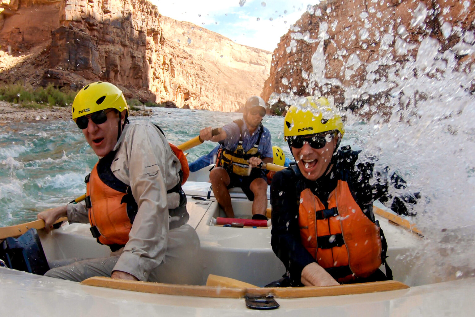 Close up image of two guests getting splashed in the front of an OARS dory while guide smiles from the oars in Grand Canyon