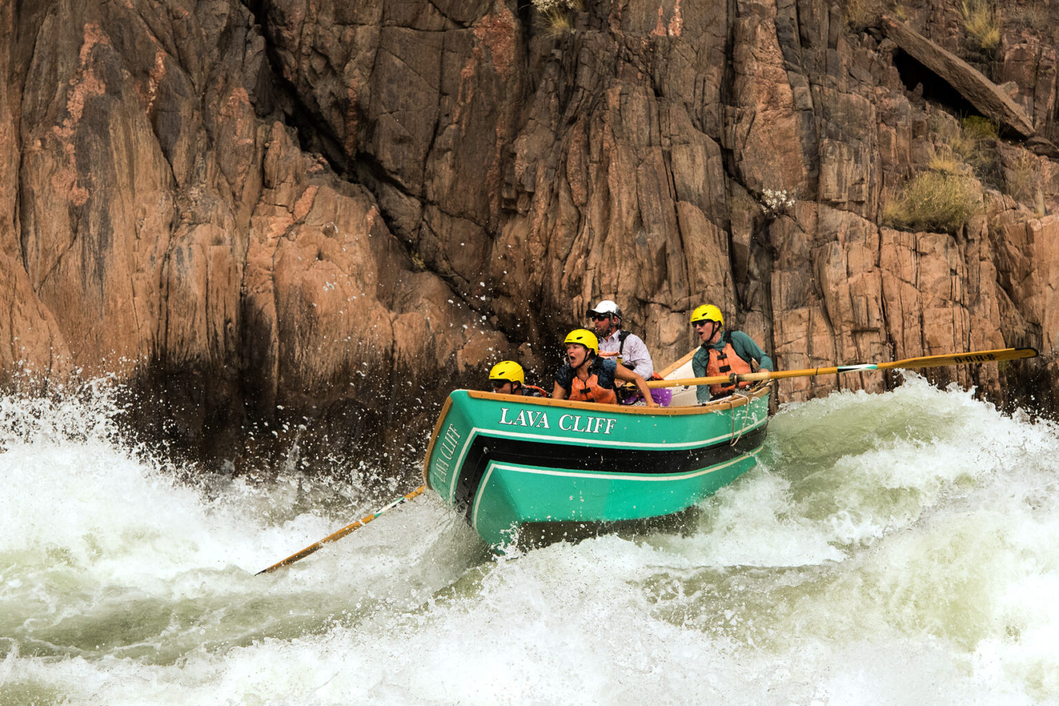 OARS guide steers a wooden dory through rough whitewater in Grand Canyon while three guests in yellow helmets hang on tight
