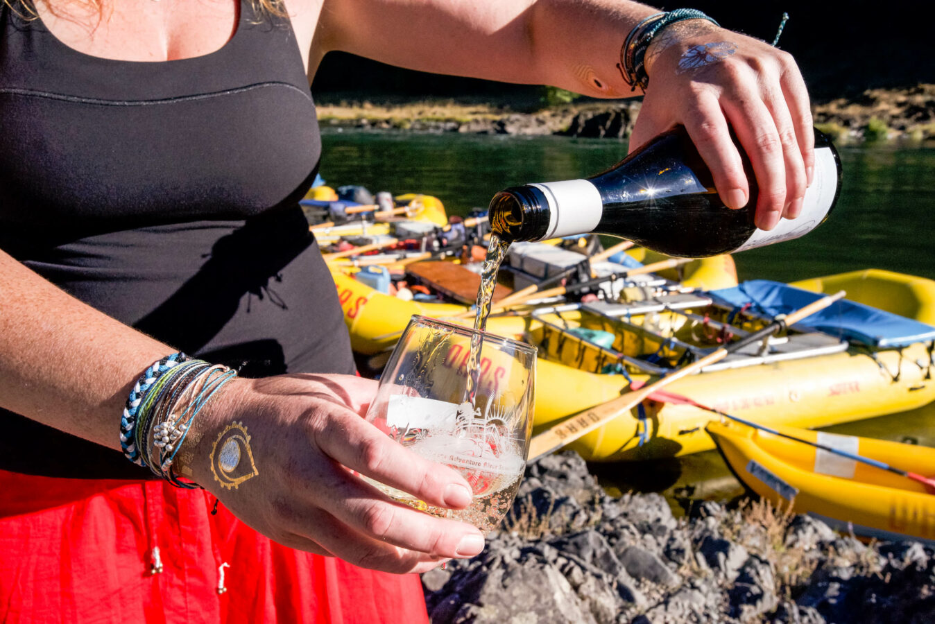 Person pouring wine into a glass on the Rogue River bank.