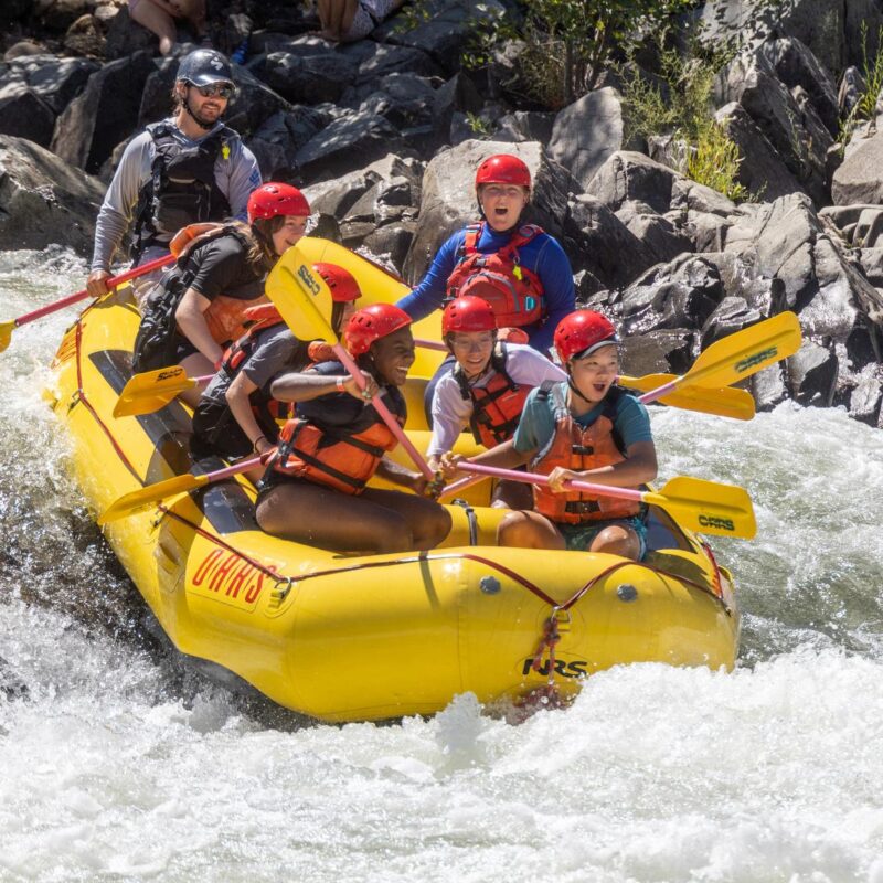A group of young people smiling while rafting through Troublemaker Rapid on the South Fork of the American River.