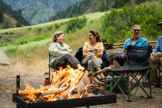 women in camp chairs chatting behind a camp fire.