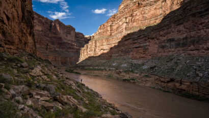 A lone person stands along the the Colorado River with the walls of Cataract Canyon towering above them